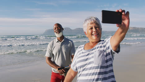 Smiling-senior-african-american-couple-walking-with-bicycles-and-taking-a-selfie-at-the-beach