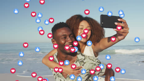 Animation-of-red-heart-love-and-like-digital-icons-over-smiling-couple-taking-selfie-on-beach