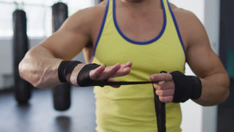 Mid-section-of-caucasian-male-boxer-wrapping-boxing-tape-on-his-hands-at-the-gym