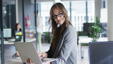 Portrait-of-caucasian-businesswoman-standing-in-office-using-laptop-looking-to-camera-and-smiling