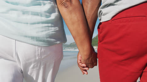 Midsection-of-senior-african-american-couple-walking-and-holding-hands-at-the-beach