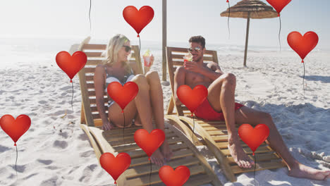 Animation-of-red-heart-love-balloons-digital-icons-over-couple-in-deckchairs-with-drinks-on-beach