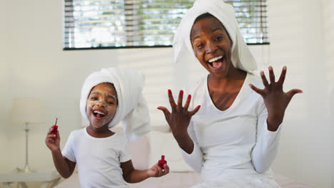 Happy-african-american-mother-and-daughter-sitting-on-bed-and-showing-painted-nails-to-camera