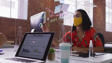 Mixed-race-businesswoman-writing-on-sneez-screen-sitting-in-front-of-computer-wearing-face-mask