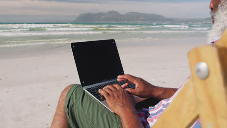 Mixed-race-senior-man-sitting-on-sunbed-and-using-a-laptop-at-the-beach