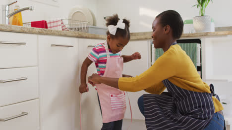 Happy-african-american-mother-and-daughter-wearing-aprons-in-kitchen