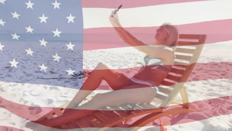 Animación-De-La-Bandera-Estadounidense-Ondeando-Sobre-Una-Mujer-En-Una-Tumbona-Tomándose-Un-Selfie-Usando-Un-Teléfono-Inteligente-En-La-Playa.