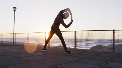 Focused-african-american-man-exercising-outdoors,-stretching-by-seaside-at-sunset