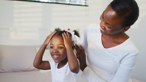 Happy-african-american-mother-and-daughter-sitting-on-bed,-mother-putting-hair-bow