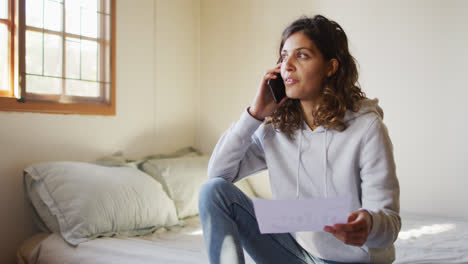 Mixed-race-woman-working-at-home,-sitting-on-bed-talking-on-smartphone-holding-paperwork-in-cottage