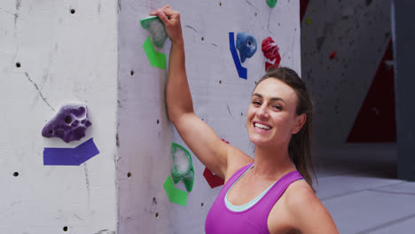 Portrait-of-smiling-caucasian-woman-grabbing-a-hold-on-a-climbing-wall-at-indoor-climbing-wall