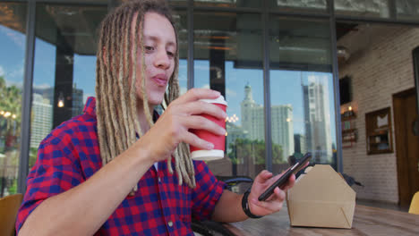 Mixed-race-man-with-dreadlocks-sitting-at-table-outside-cafe-drinking-coffee-and-using-smartphone