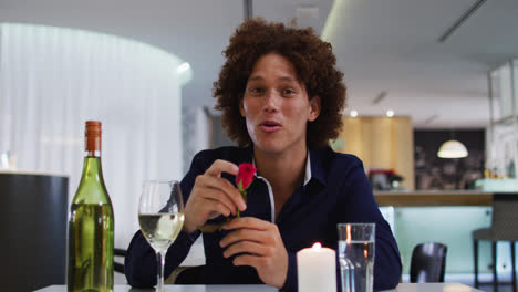 Mixed-race-man-having-romantic-dinner-at-restaurant-holding-red-rose-and-talking-to-camera