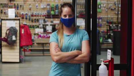 Caucasian-female-shopkeeper-wearing-face-mask-in-the-doorway-of-sports-shop-with-arms-crossed