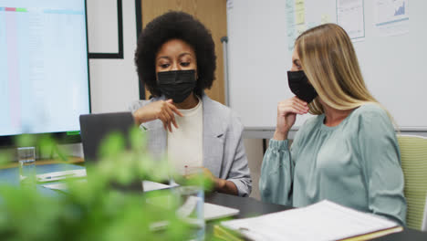 Two-diverse-businesswomen-wearing-face-masks-working-together-using-laptop-at-desk-in-office