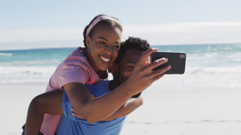 Pareja-Afroamericana-Tomando-Selfie-Con-Teléfono-Inteligente,-Hombre-Llevando-A-Una-Mujer-A-Cuestas-En-La-Playa
