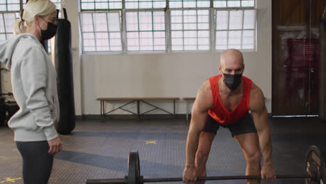Caucasian-muscular-man-wearing-face-mask-and-lifting-weight-bar-with-coach