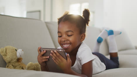 Glückliches-Afroamerikanisches-Mädchen,-Das-Zu-Hause-Mit-Einem-Digitalen-Tablet-Auf-Dem-Sofa-Liegt