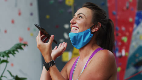 Lachende-Kaukasische-Frau-Mit-Gesichtsmaske,-Die-Ihr-Smartphone-An-Einer-Indoor-Kletterwand-Benutzt