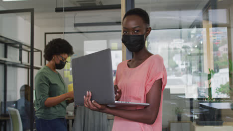 Happy-african-american-businesswoman-wearing-face-mask-using-laptop,-looking-to-camera-in-office