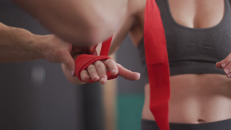 Mid-section-of-male-trainer-wrapping-boxing-tape-on-hands-of-female-boxer-at-the-gym