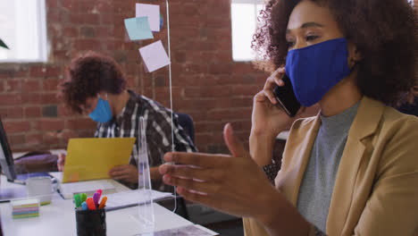 Mixed-race-businesswoman-talking-on-phone-sitting-in-front-of-computer-wearing-face-mask