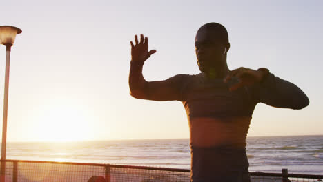 Fokussierter-Afroamerikanischer-Mann,-Der-Sich-Dehnt-Und-Bei-Sonnenuntergang-Im-Freien-Am-Meer-Trainiert