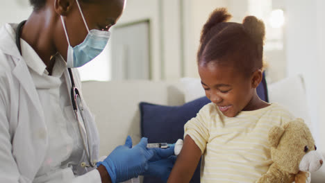Afroamerikanische-Ärztin-Mit-Gesichtsmaske,-Die-Lächelndem-Mädchen-Zu-Hause-Eine-Covid-19-Impfung-Verabreicht