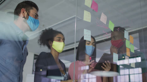 Diverse-colleagues-wearing-face-masks-using-memo-notes-on-glass-wall-having-a-discussion