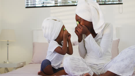 Happy-african-american-mother-and-daughter-wearing-towels-sitting-on-bed-putting-cucumbers-on-eyes