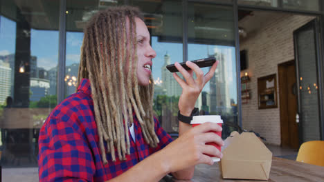 Mixed-race-man-with-dreadlocks-sitting-at-table-outside-cafe-drinking-coffee-and-using-smartphone