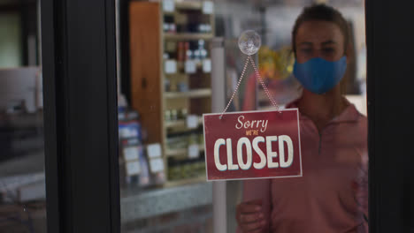 Caucasian-female-shopkeeper-wearing-face-mask-changing-closed-to-open-sign-in-window-of-sports-shop