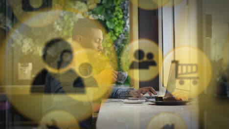 Múltiples-íconos-Digitales-Contra-Un-Hombre-Afroamericano-Tomando-Café-Mientras-Usa-Una-Computadora-Portátil-En-Una-Cafetería