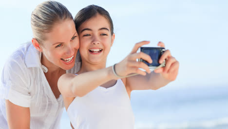 Portrait-of-smiling-caucasian-mother-and-daughter-on-holiday-taking-photos-by-sea