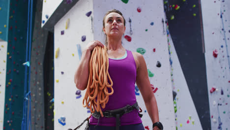 Caucasian-woman-with-rope-over-her-shoulder-preparing-for-climb-at-indoor-climbing-wall