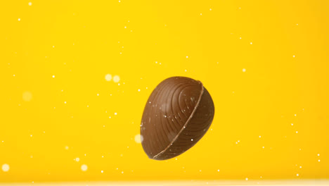 Animation-of-white-spots-floating-over-chocolate-easter-egg-falling-and-bouncing,-on-yellow