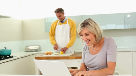 Woman-using-laptop-while-her-husband-is-slicing-vegetables