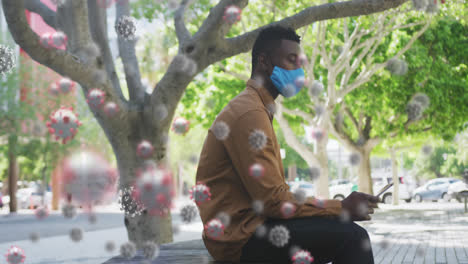 Multiple-covid-19-cells-floating-against-african-american-man-wearing-face-mask-using-smartphone