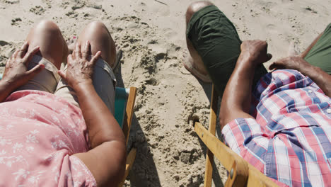 Senior-african-american-couple-sitting-on-sunbeds-and-holding-hands-at-the-beach