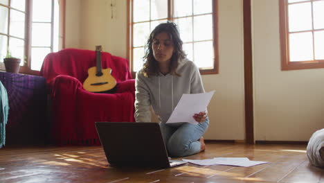 Mixed-race-woman-working-at-home,-kneeling-on-floor-using-laptop-and-holding-paperwork-in-cottage
