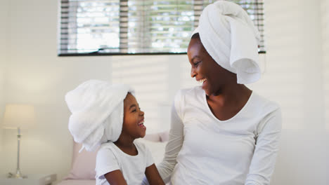 African-american-mother-and-daughter-wearing-towels-sitting-on-bed-and-laughing