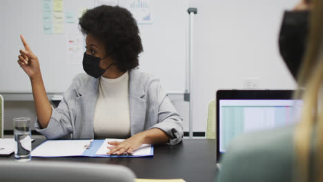 African-american-businesswoman-wearing-face-mask-sitting-at-desk-talking-to-colleagues-in-office