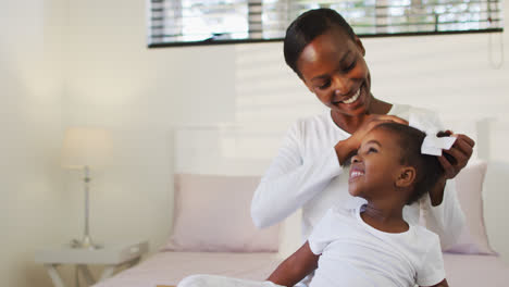 Happy-african-american-mother-and-daughter-sitting-on-bed,-mother-putting-hair-bow