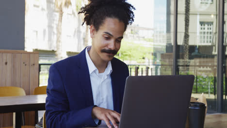 Mixed-race-man-with-moustache-sitting-at-table-outside-cafe-using-laptop