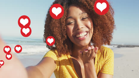Animation-of-red-heart-love-digital-icons-over-smiling-woman-blowing-kisses-on-beach