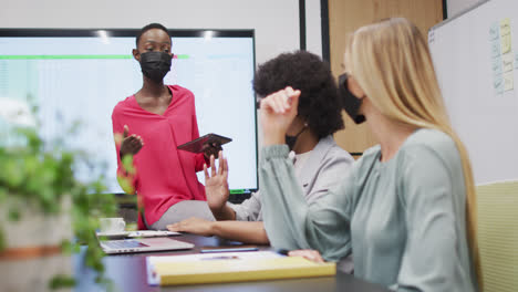 Three-diverse-businesswomen-wearing-face-masks-in-discussion-at-office-meeting,-one-holding-tablet