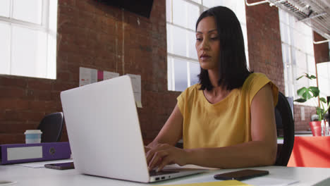 Mixed-race-businesswoman-sitting-at-desk-using-laptop-looking-ahead