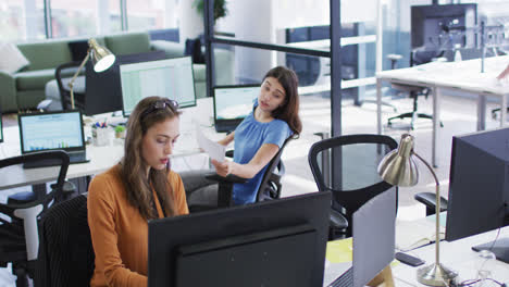Two-caucasian-businesswomen-sitting-at-desks-using-computers-turning-around-to-talk-to-each-other