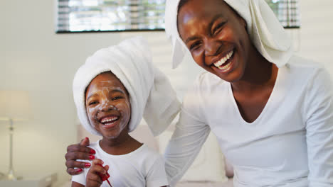 Happy-african-american-mother-and-daughter-wearing-towels-sitting-on-bed-and-looking-to-camera