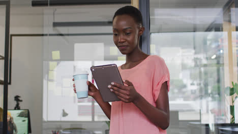 African-american-businesswoman-drinking-takeaway-coffee-using-tablet-and-smiling-to-camera-in-office